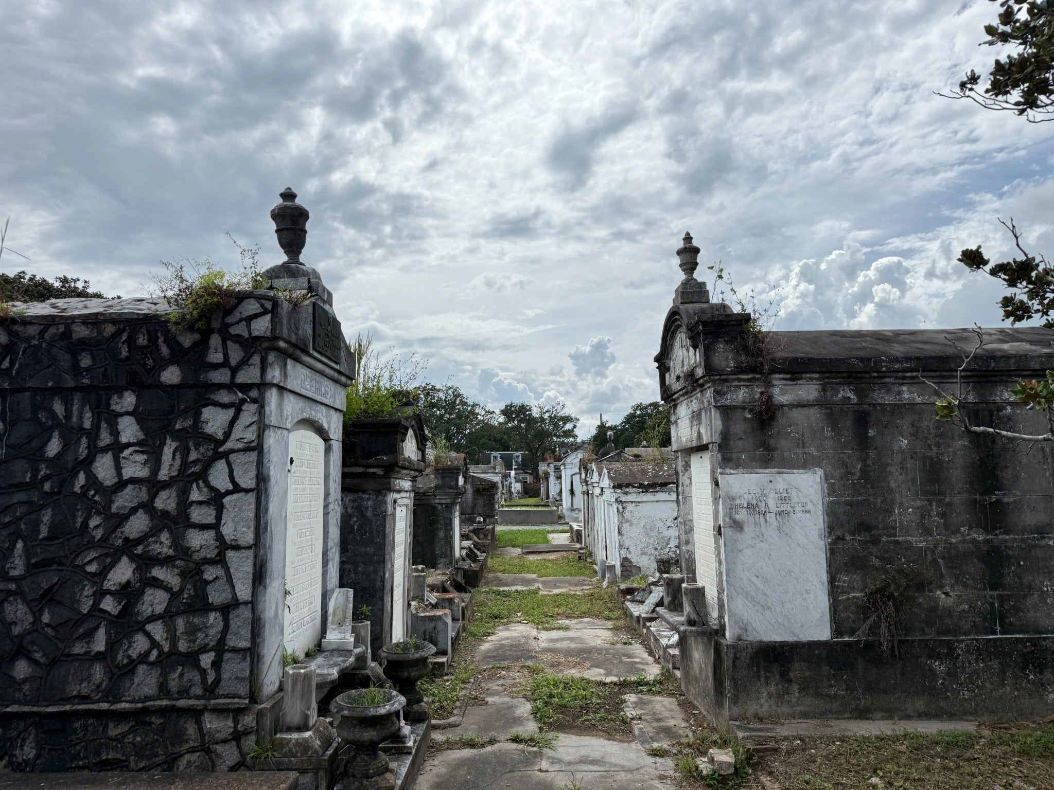 A narrow pathway lined with weathered above-ground tombs in Lafayette Cemetery No. 1, with ornate stone mausoleums, carved memorials, and urns surrounded by grass and vegetation beneath a cloudy sky.