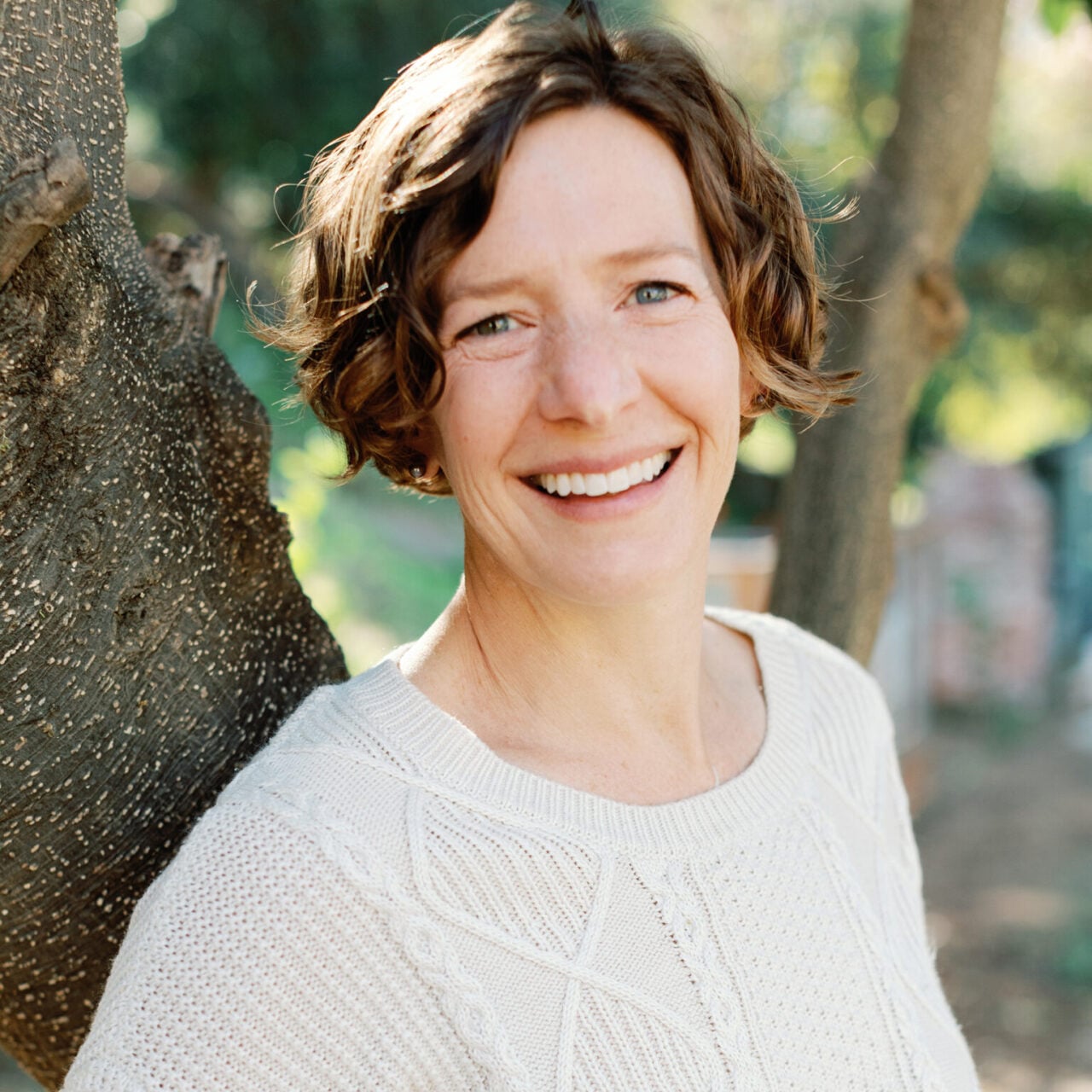 Nicola Twilley, author and journalist, smiling outdoors in a cream sweater, leaning against a tree