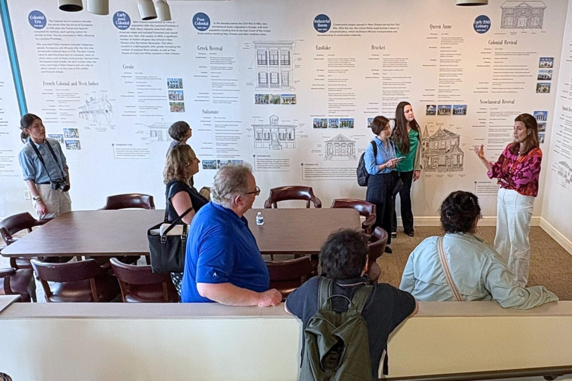 A guide gives a presentation to a small group inside an educational exhibit space, standing in front of a large wall display illustrating and explaining historic New Orleans architectural styles.