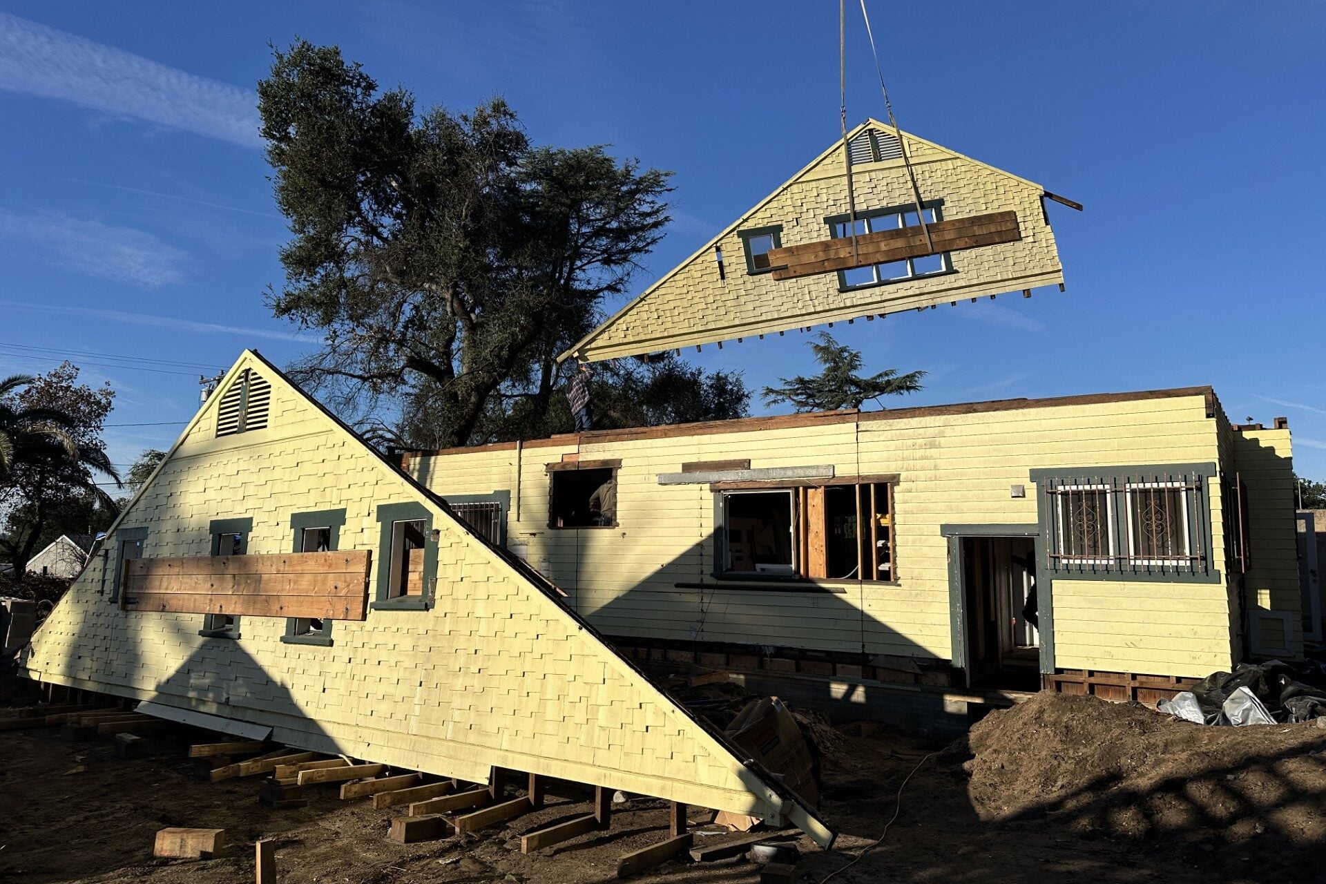 A yellow wood-frame house cut in half, with a matching gable-end section suspended by crane above the main structure, both sections braced with diagonal wood beams across the window openings; the main house body rests on temporary wood cribbing supports.