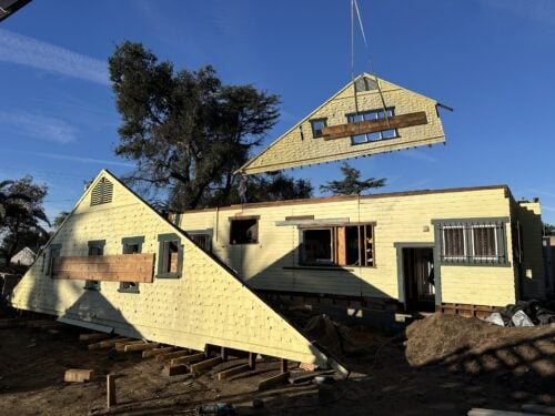A yellow wood-frame house cut in half, with a matching gable-end section suspended by crane above the main structure, both sections braced with diagonal wood beams across the window openings; the main house body rests on temporary wood cribbing supports.