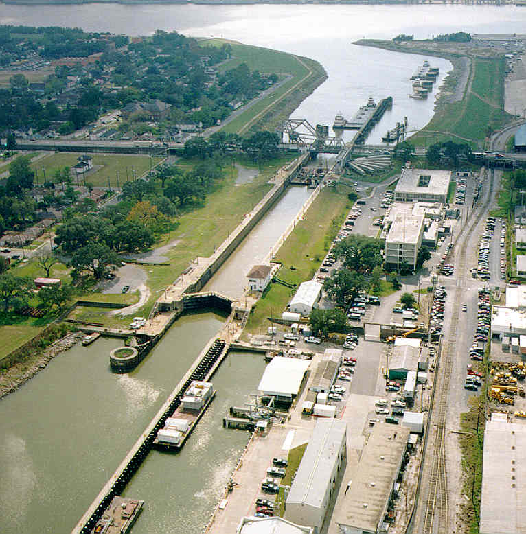 Undated aerial photograph of the Industrial Canal Lock in New Orleans before Hurricane Katrina, showing barges and towboats navigating the waterway near the St. Claude Avenue Bridge. Industrial buildings, rail lines, and roadways line the canal, with the Lower Ninth Ward to the left and Bywater to the right.