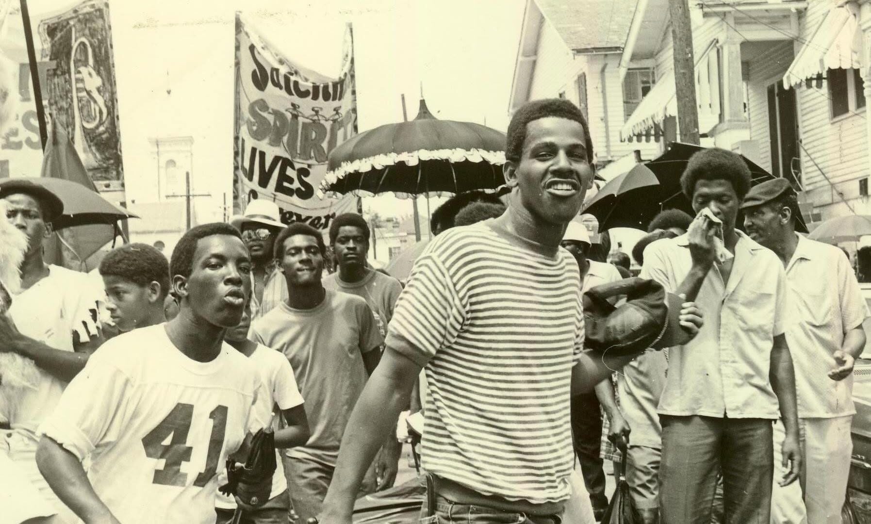 A black-and-white photograph of a second line honoring Louis Armstrong on July 10, 1971, passing in front of Blandin Funeral Home in New Orleans. A smiling man in a striped shirt walks among a crowd of mourners and participants carrying umbrellas and banners. Photograph by an unknown photographer; image shared by Yoshio Toyama in 2020.