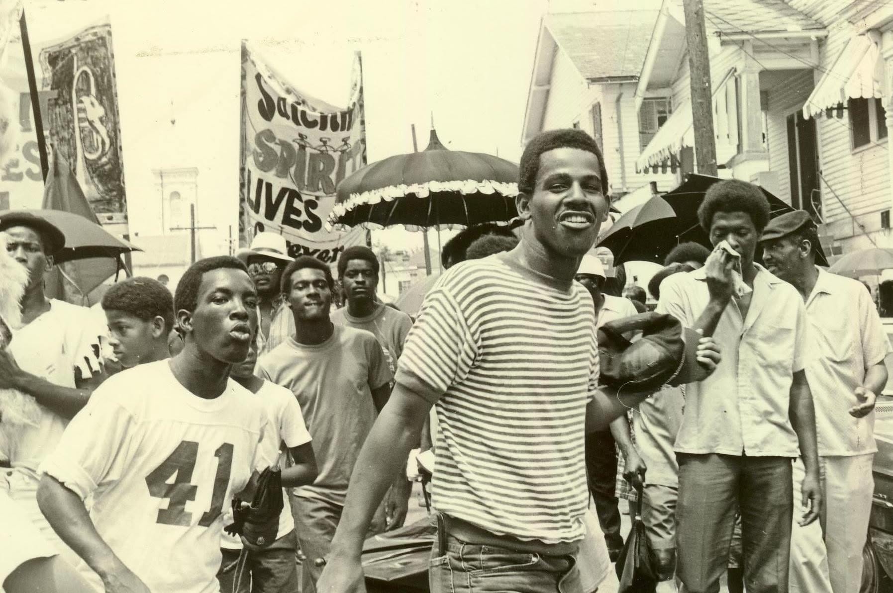 A black-and-white photograph of a second line honoring Louis Armstrong on July 10, 1971, passing in front of Blandin Funeral Home in New Orleans. A smiling man in a striped shirt walks among a crowd of mourners and participants carrying umbrellas and banners. Photograph by an unknown photographer; image shared by Yoshio Toyama in 2020.