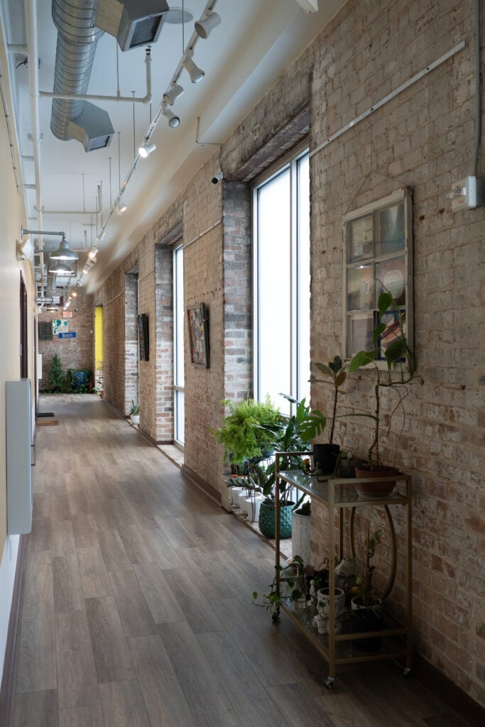 Brick-lined hallway at St. Joe Lofts with tall windows, exposed ductwork and houseplants