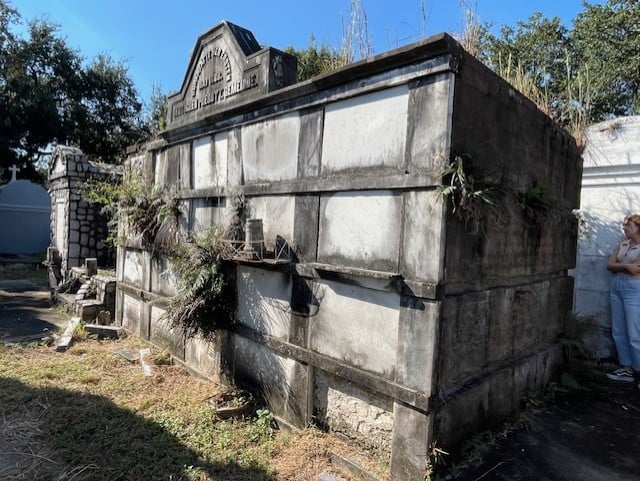 German Presbyterian tomb in Lafayette Cemetery No. 1, showing its weathered marble burial panels, darkened stonework, and ornate pediment inscription. Vegetation grows along the base and sides of the tomb.
