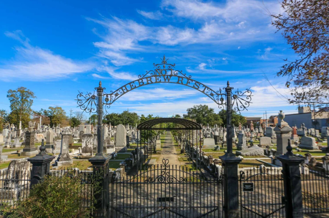 Ornate wrought-iron entrance gate marked “Hebrew Rest” at a New Orleans cemetery, with rows of headstones and monuments lining a central pathway beneath a covered metal trellis. The cemetery is framed by trees and a bright blue sky.
