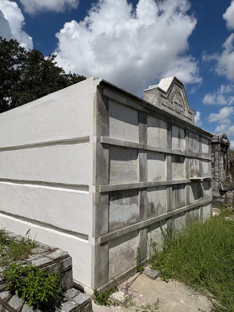 Rear and side view of the German Presbyterian tomb in Lafayette Cemetery No. 1, with weathered burial panels, a newly stuccoed white wall, and tall grass surrounding the structure under a bright blue sky.