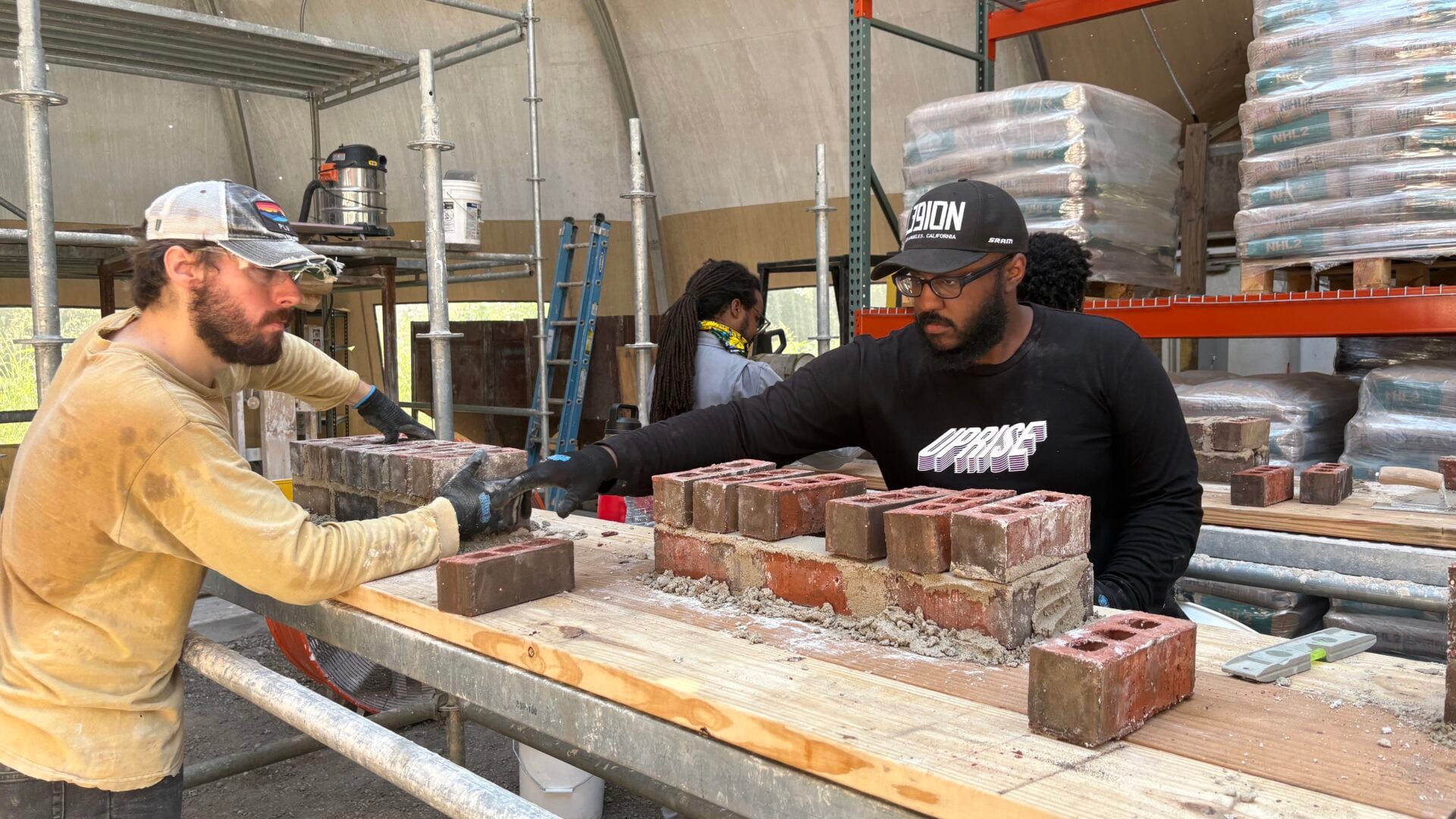 Two workers build a brick wall on a wooden workbench inside a covered construction area, with scaffolding, tools, ladders, and stacks of building materials surrounding them. Both workers wear protective gloves as they handle bricks and mortar.