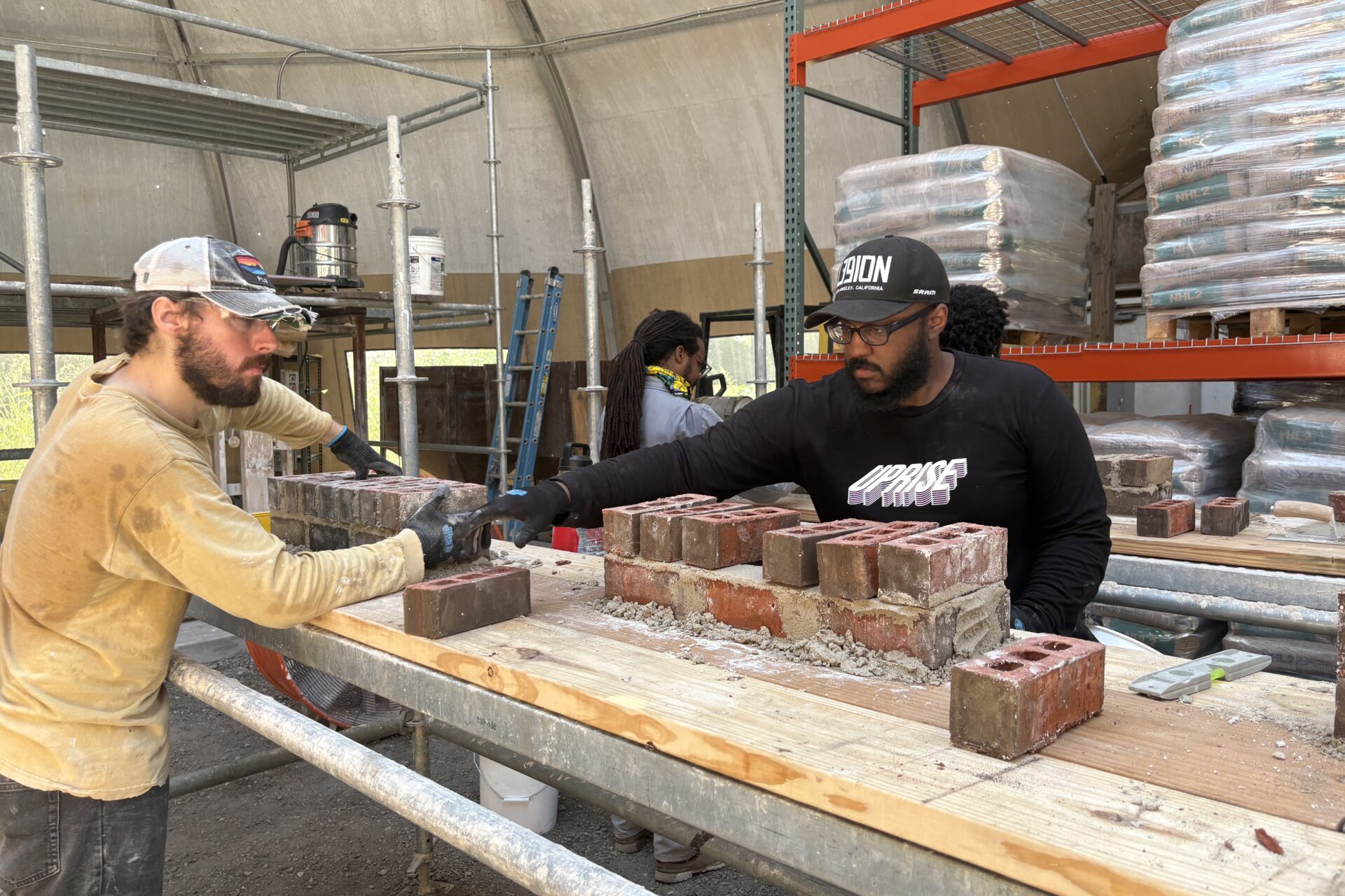 Two workers build a brick wall on a wooden workbench inside a covered construction area, with scaffolding, tools, ladders, and stacks of building materials surrounding them. Both workers wear protective gloves as they handle bricks and mortar.