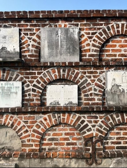 Historic brick wall vault in a New Orleans cemetery, featuring multiple marble burial plaques set into decorative brick arches.